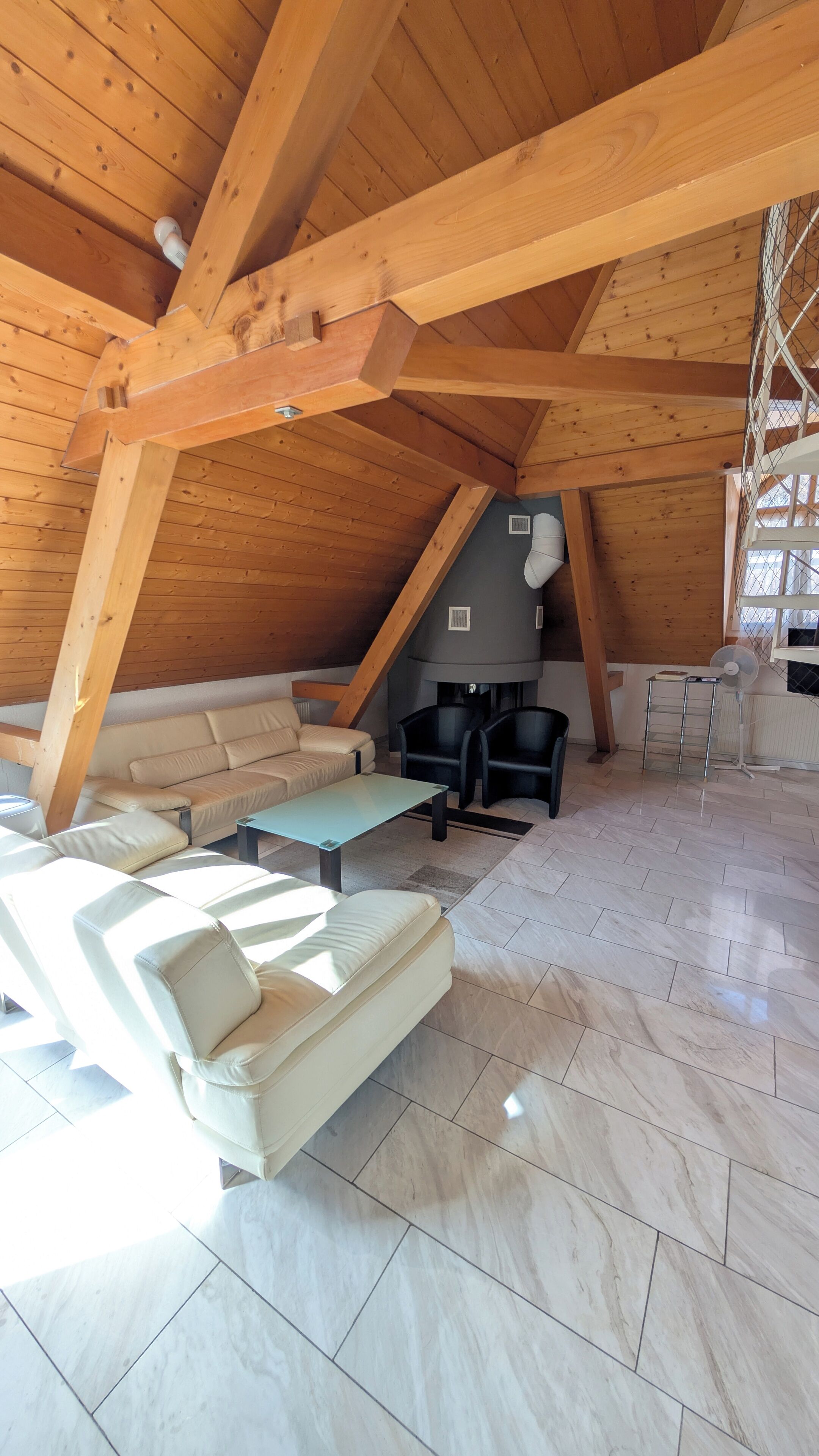 Amplia habitación en el ático con techo de madera alto, un sofá color beige, una mesita y una chimenea.