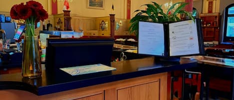 Un mostrador de recepción con un cartel que dice 'RECEPTION', un jarrón con flores rojas, una pizarra de menú y un monitor de computadora.
