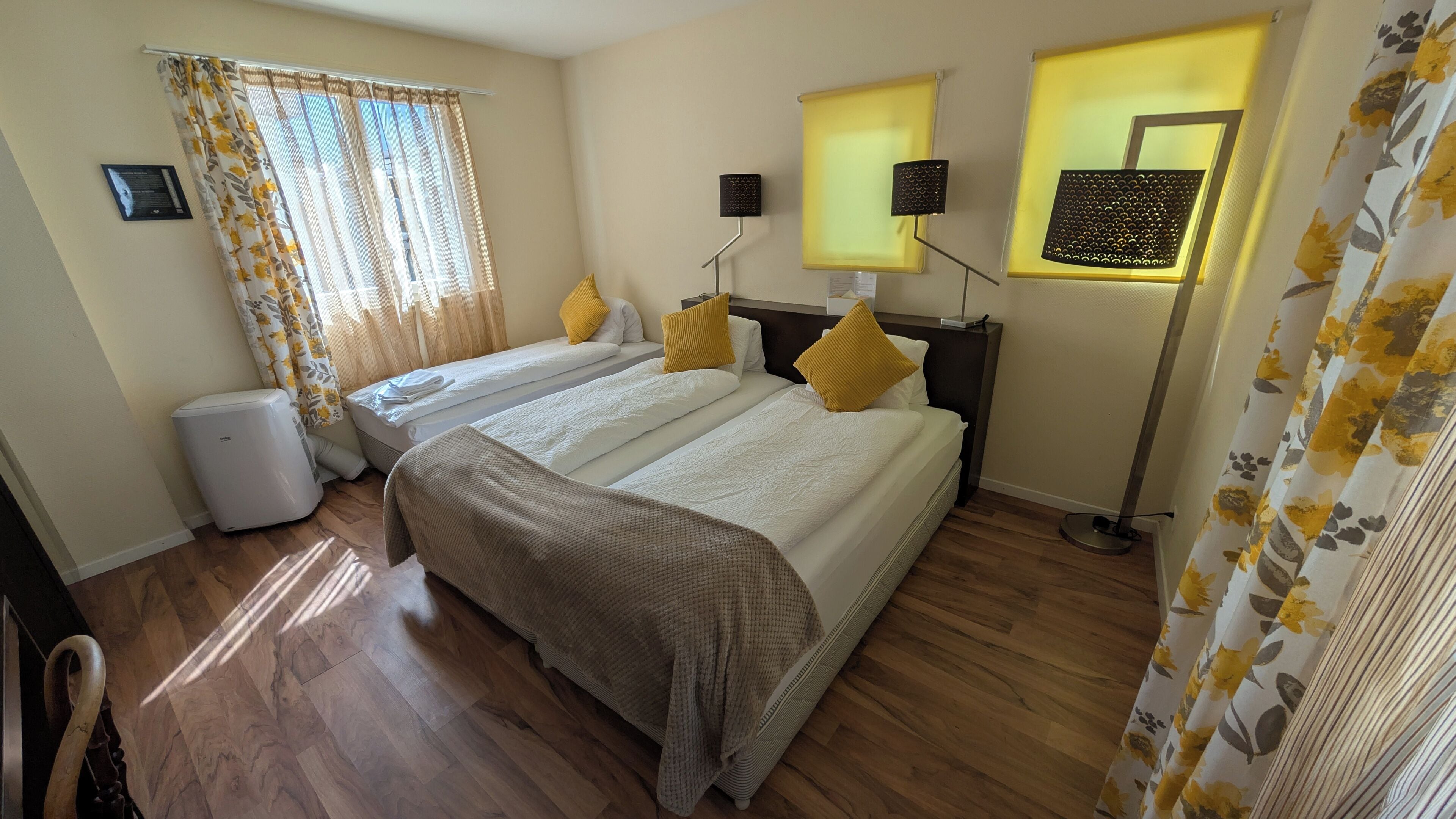 A hotel room with two beds, a large window with curtains, a standing lamp, and a small air conditioner.