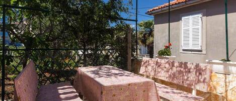 Un patio al aire libre con una mesa y bancos cubiertos con manteles a cuadros, rodeado por una valla de metal y árboles.