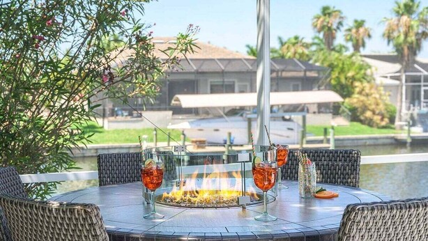 A round outdoor dining table with wicker chairs, a fire pit in the center, and drinks on the table.