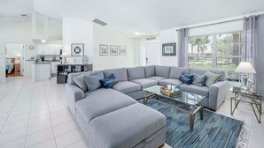 A modern living room with a grey sectional sofa, a glass coffee table, and a view of the kitchen and dining area in the background.