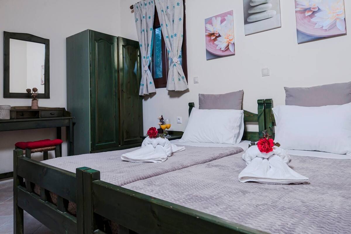 A neatly made bed with towels and flowers, a wooden wardrobe, a desk with a mirror, and a wall with framed pictures.
