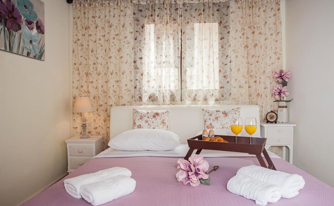 A neatly made bed with white linens, a tray of orange juice, and a vase of flowers on a purple bedspread.