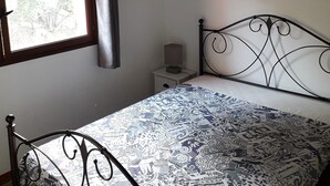 A bedroom with a metal bed frame, a patterned bedspread, a nightstand with a lamp, a window with curtains, and a cityscape painting on the wall. - T4 plus T2 independent. (Pietrosella)