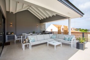 Patio esterno con divano bianco, tavolino da caffè e tavolo da pranzo con sedie. Si trova un barbecue e una pianta in vaso sulla terrazza.
