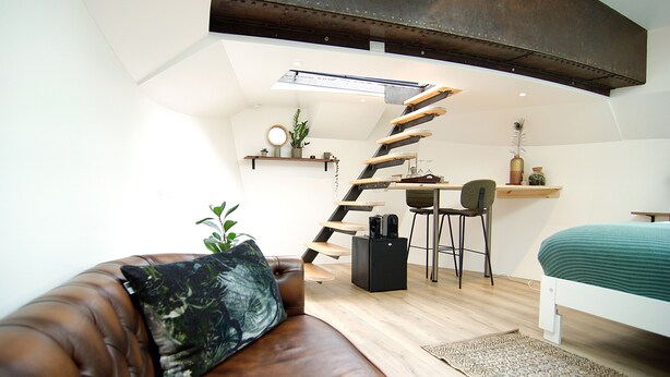 Camera da letto moderna mansardata con soffitto spiovente, scala a chiocciola, divano in pelle marrone, letto verde e una piccola area scrivania.