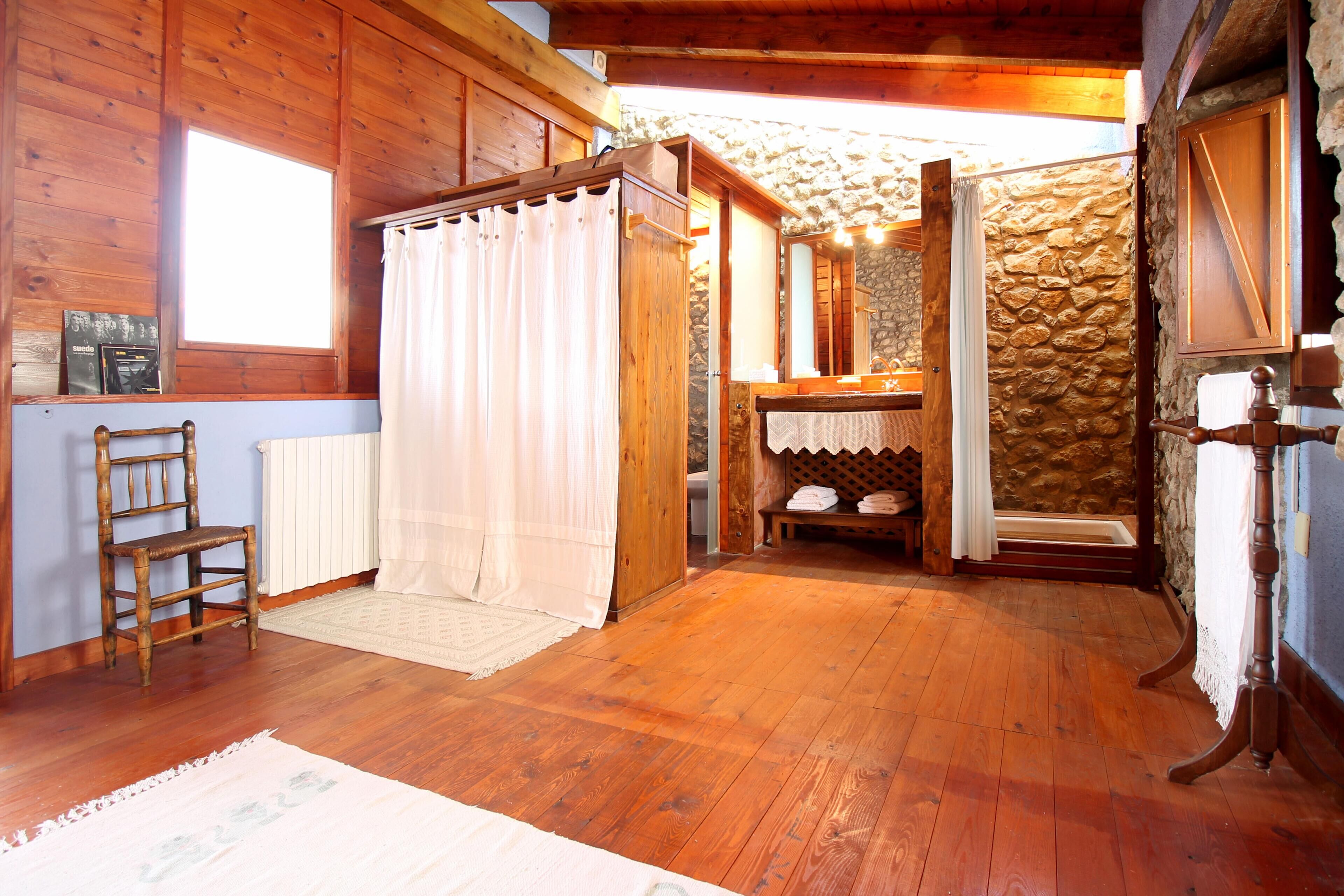 Interior de una cabaña de madera con una silla de madera, una cortina blanca, un muro de piedra y una mesa de madera.