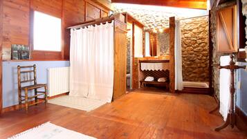 Interior de una cabaña de madera con una silla de madera, una cortina blanca, un muro de piedra y una mesa de madera.