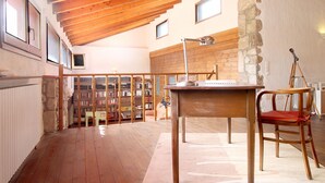 A spacious room with wooden flooring, a high ceiling with exposed beams, a desk, a chair, a tripod, and a bookshelf filled with books.
