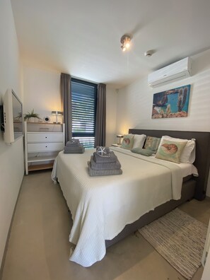 A hotel room with a bed, bedside tables, a desk, a chair, a window with curtains, an air conditioning unit, and a wall-mounted artwork.