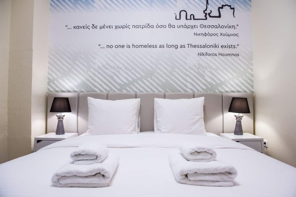 Ein Hotelzimmer mit einem großen Bett, zwei Nachttischen mit Lampen und einer Wand mit einem Zitat in Griechisch.