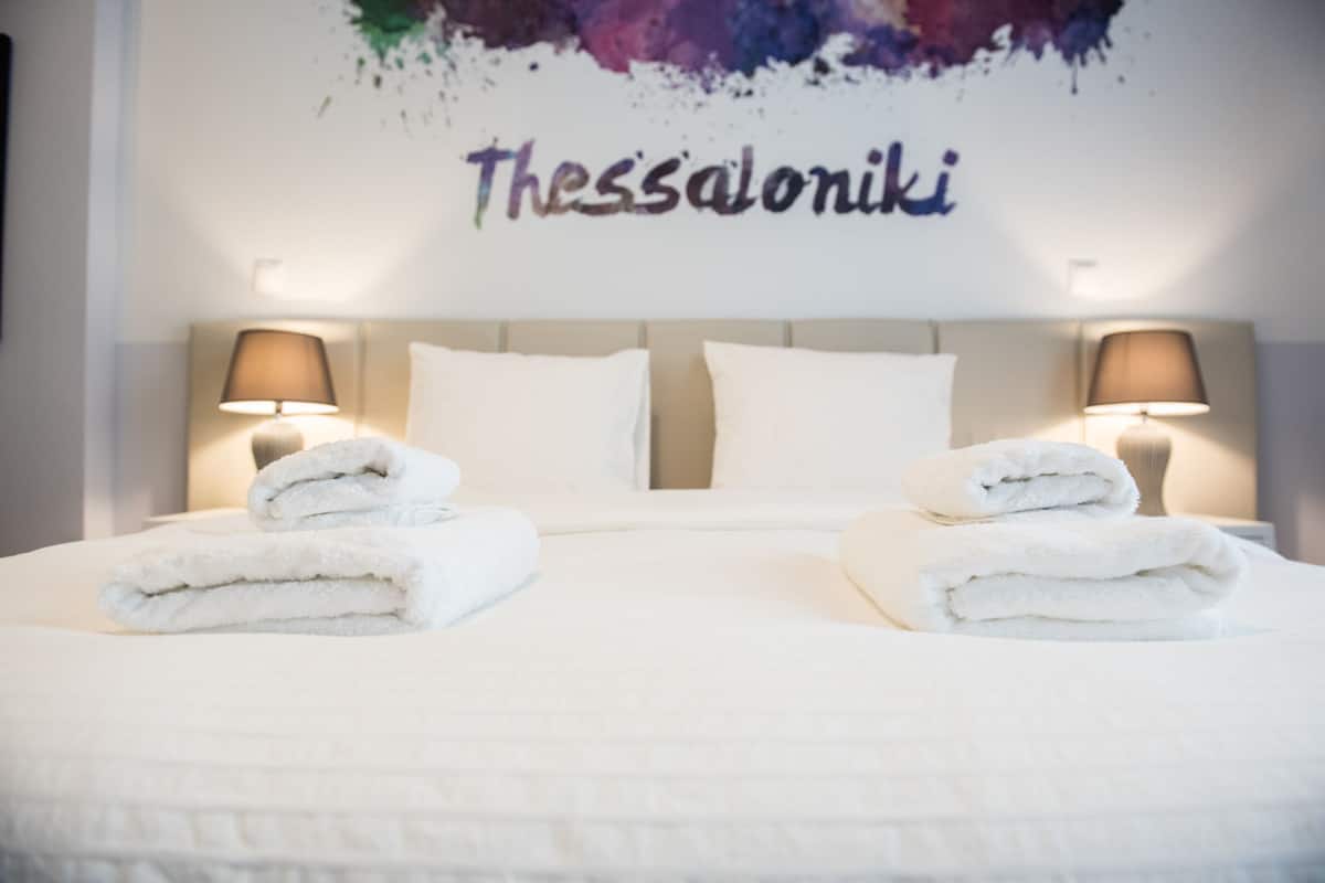 Ein ordentlich bezogenes Bett mit zwei gefalteten Handtüchern, einem Kopfteil mit zwei Wandlampen und einer Wand, auf der in einer stilisierten Schriftart "Thessaloniki" steht.