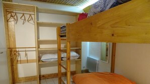 Un lit superposé avec un oreiller orange, une échelle en bois et une étagère où sont suspendus des vêtements et des sacs.