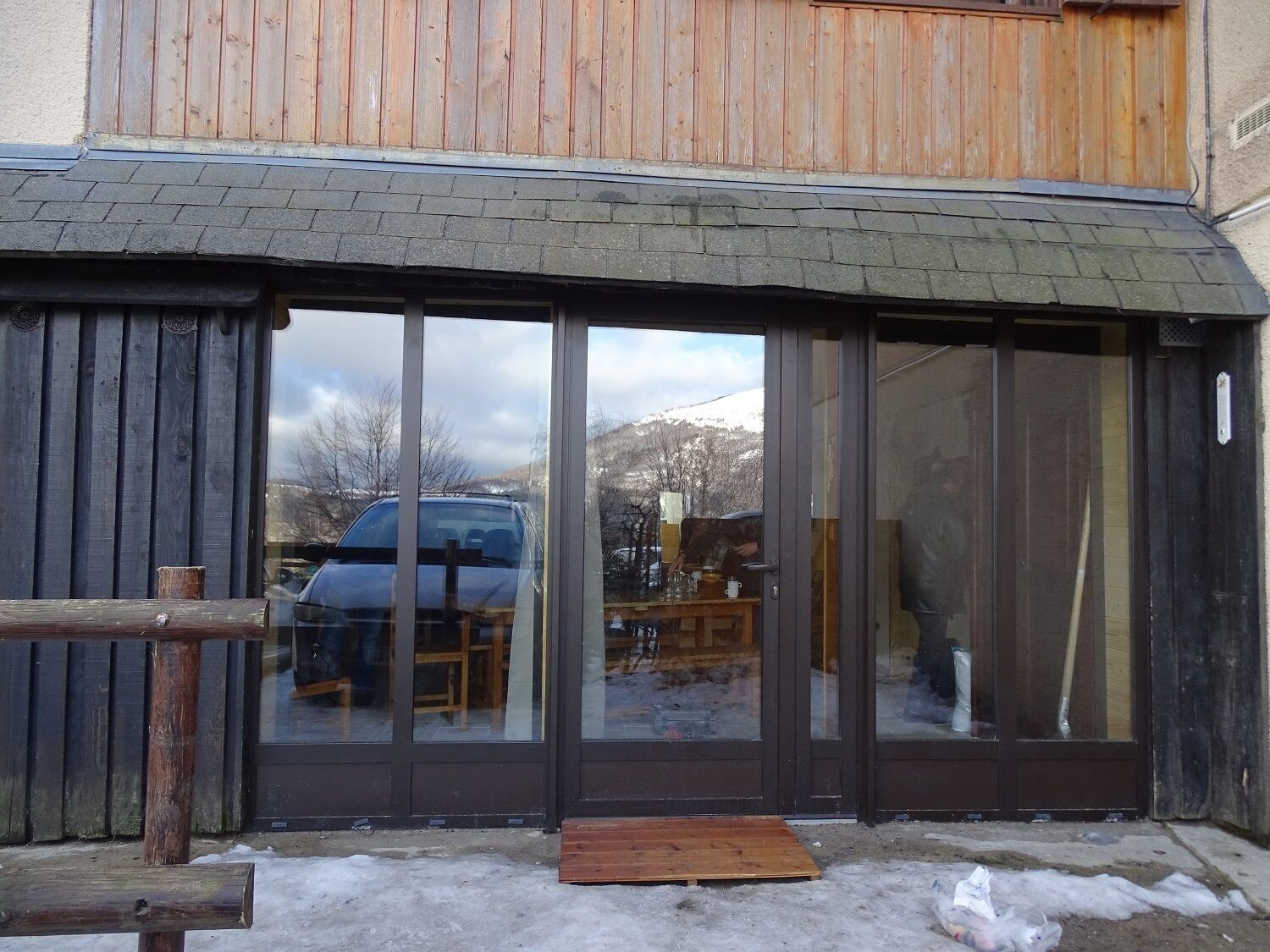 Un bâtiment doté de grandes portes vitrées, d’une terrasse en bois et d’une voiture gar ée à l’extérieur.