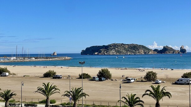 Una playa con una isla rocosa al fondo, un puerto deportivo con veleros y varios vehículos estacionados.