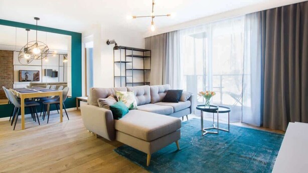 A modern living room with a sofa, ottoman, dining table, and chairs. There is a large window with curtains, a chandelier, and a small side table with flowers. - Modern Apartment | Parking, Balcony with View (Gdansk)