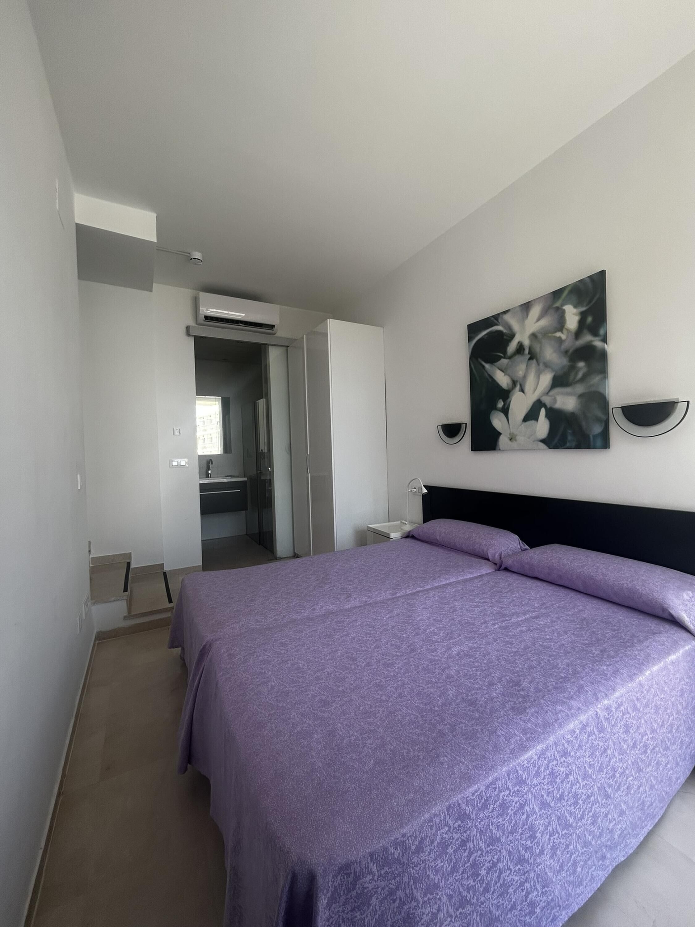 A bedroom with a bed, a nightstand, a wall-mounted lamp, and a painting.