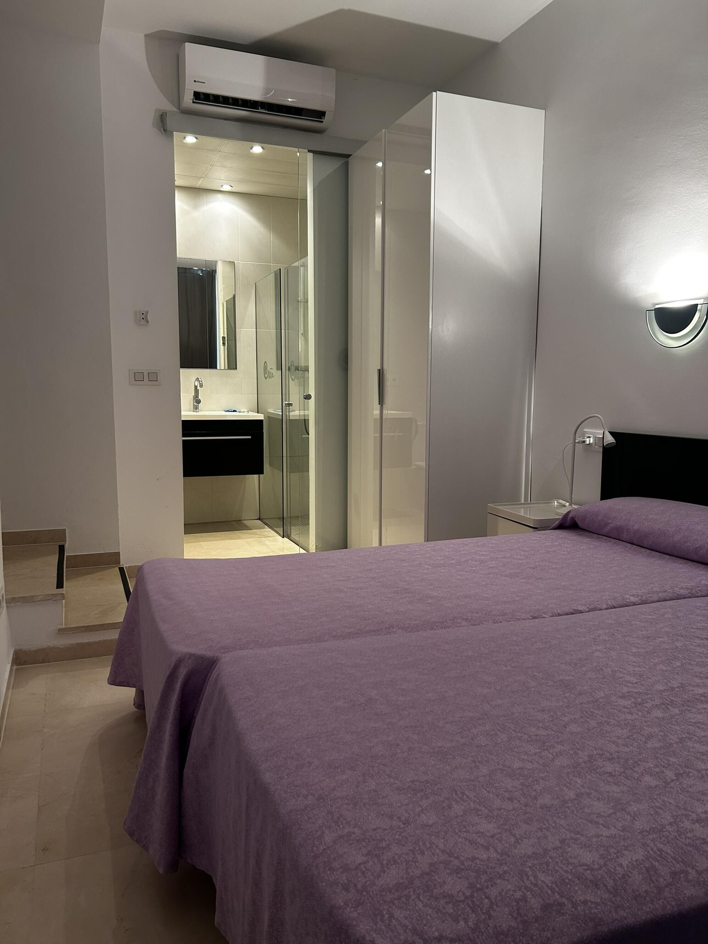 A bedroom with a bed, a wardrobe, a bathroom with a glass shower enclosure, and an air conditioning unit.