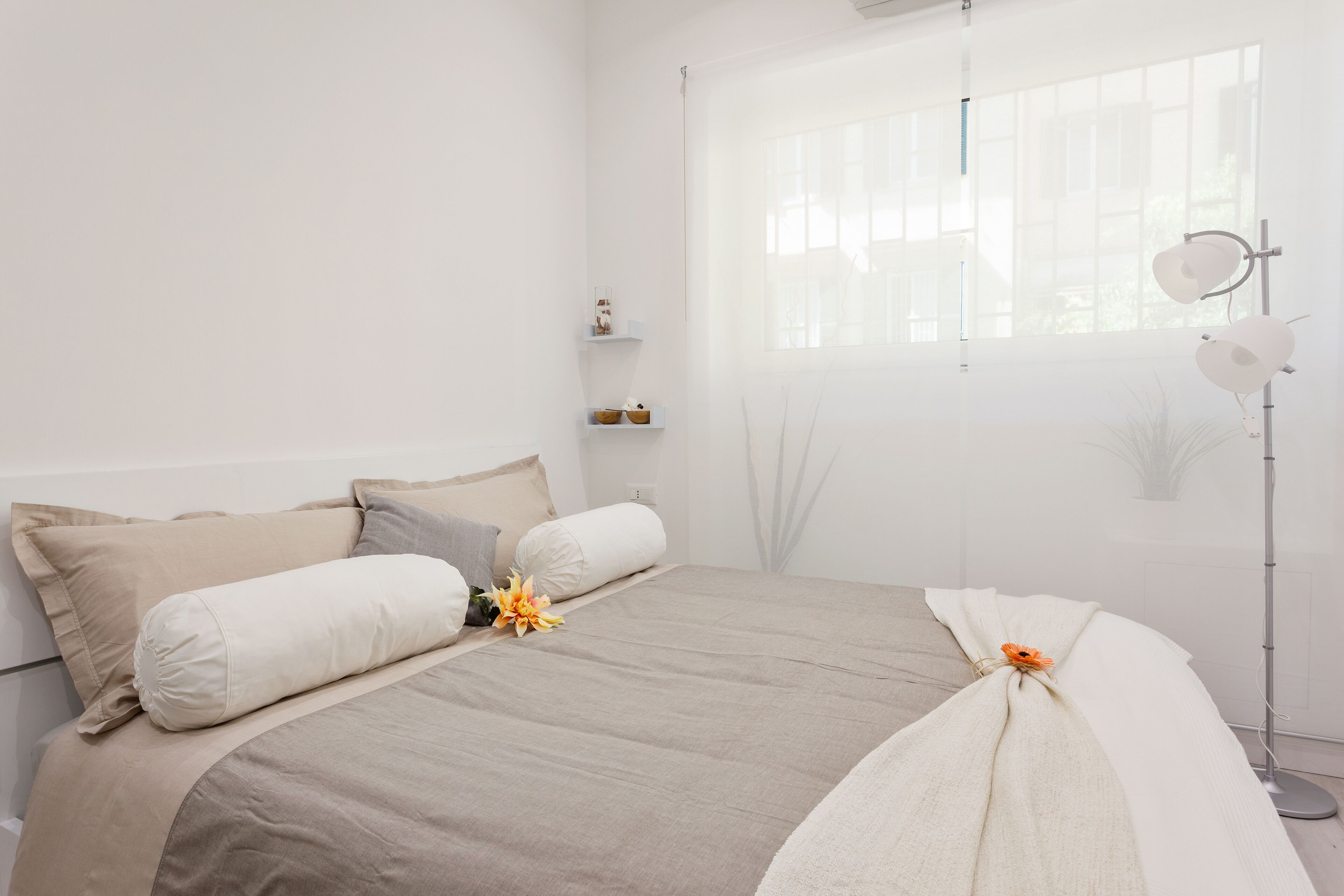 A minimalist bedroom with a large bed, a bedside table, a floor lamp, and a window with blinds.