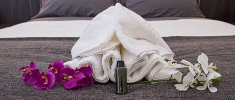 A neatly made bed with a white towel, purple and white flowers, and a bottle on a gray bedspread.