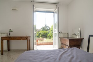 Quarto com cama, escrivaninha, cômoda e porta de vidro deslizante para uma varanda.