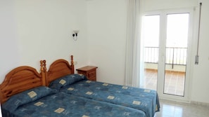 A bedroom with a wooden bed, a small bedside table, a wall-mounted light, and a sliding glass door to a balcony. - Apartaments  Nàutic Xaloc first line of the sea (Escala, L')