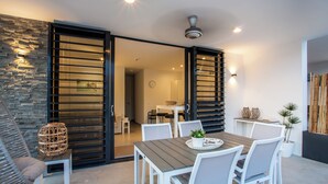 A modern dining area with a wooden table and white chairs, a stone wall, and a sliding glass door leading to a bathroom. - Hasta La Vista Royal – Tropical 7BR Villa for Groups, Jan Thiel (Jan Thiel)