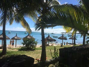 Eine Strandansicht mit Palmen, Reetdachschirmen und Booten auf dem Wasser.