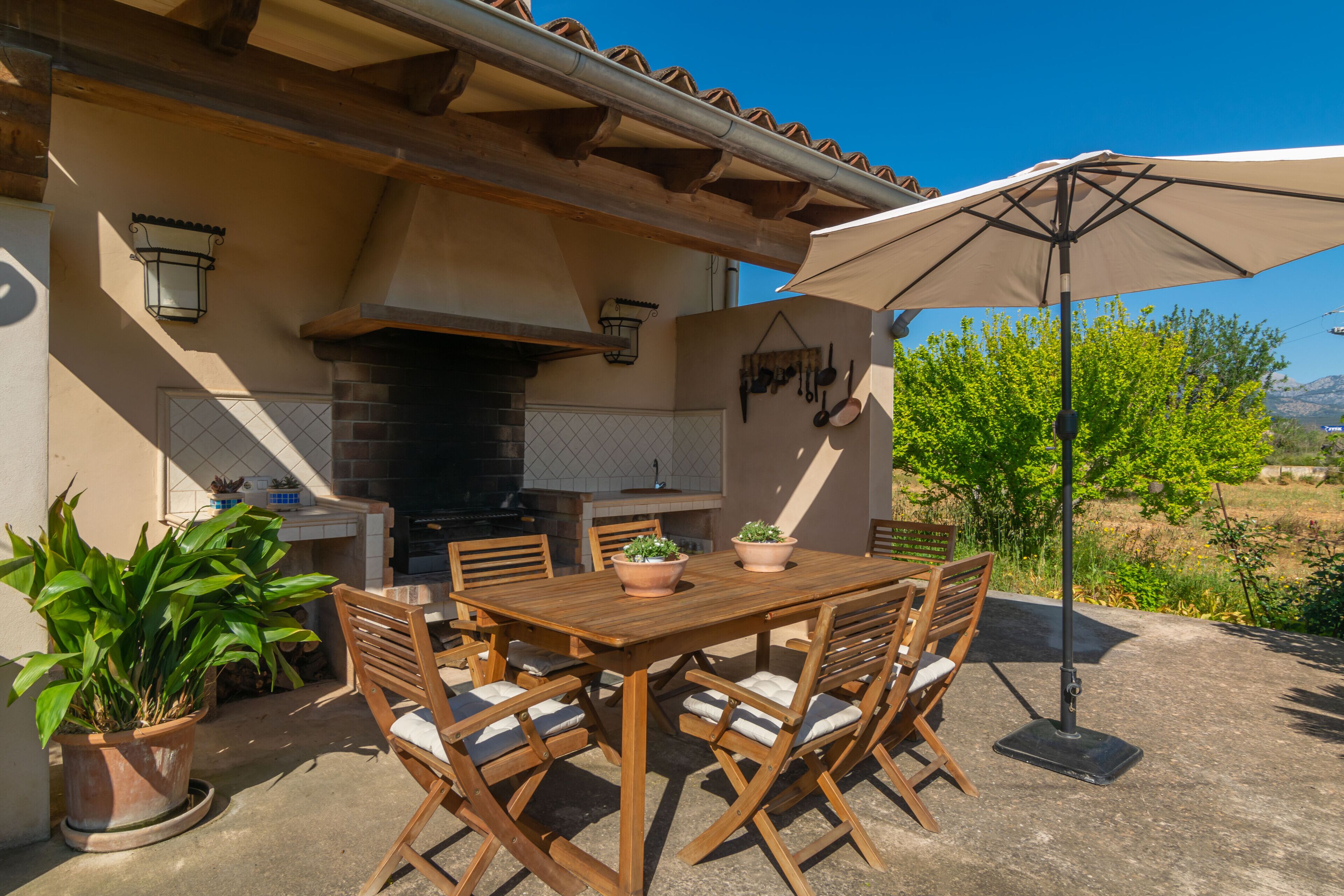 Un patio exterior con mesa y sillas de madera, chimenea de ladrillo y plantas en macetas.