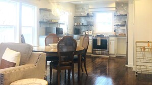 A spacious living area with a dining table, chairs, and a sofa. A kitchen is visible in the background with modern appliances and a stone backsplash.
