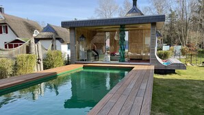 Une piscine entourée d’une terrasse en bois, un petit kiosque avec une structure en verre et, au fond, une maison au toit de chaume.