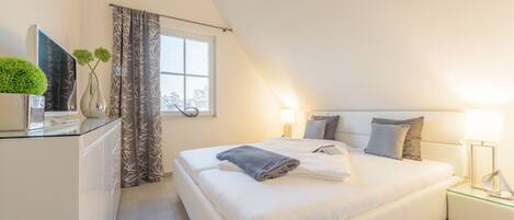 Une chambre moderne avec un grand lit, des lampes de chevet, une table en verre agrémentée d’objets décoratifs et une fenêtre ornée de rideaux à motifs.