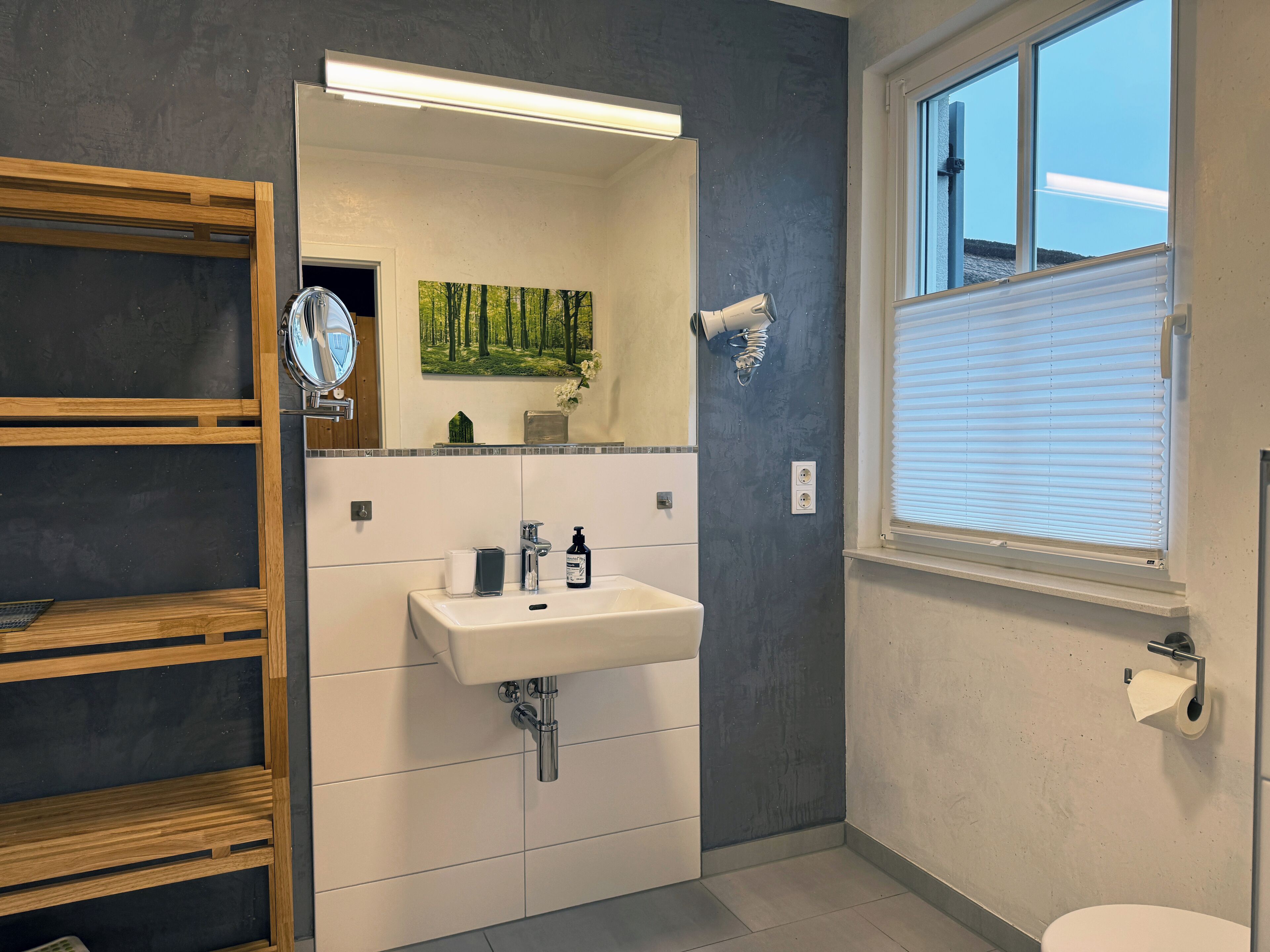 Une salle de bain moderne avec une étagère en bois, un lavabo avec un miroir, un tableau représentant une forêt et une fenêtre avec des stores.
