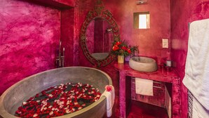 A bathroom with a large bathtub filled with rose petals, a mirror, and a sink with a vase of flowers.