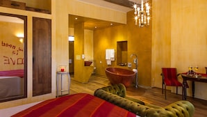 A modern hotel room with a large bed, a wooden bathtub, a red chair, and a dining area with two glasses of wine.