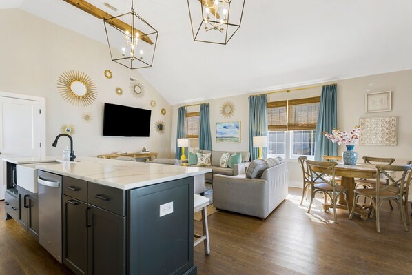 A modern kitchen with a central island, a dining area with a wooden table and chairs, and a living room with a sofa and a TV. - Guesthouse Charleston: WEST (62 & 64) (Charleston)