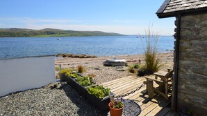 A wooden deck with a stone building, a garden with plants, and a view of a lake with boats.