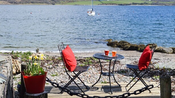 A lakeside seating area with a table and chairs, a potted plant, and a sailboat in the distance.
