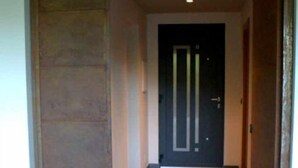 A hallway with a dark door at the end, a light-colored wall, and a brown floor.