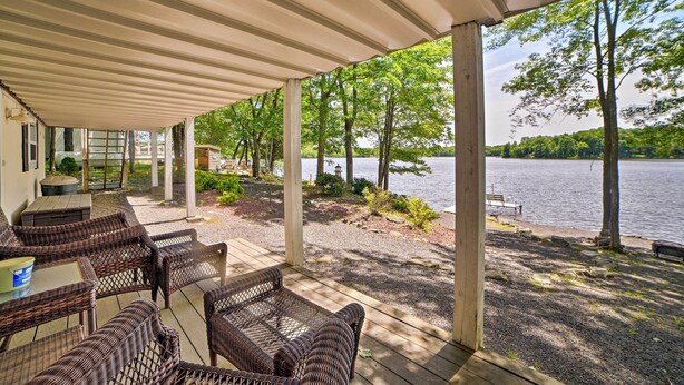A covered outdoor seating area with wicker furniture, a table, and a view of a lake surrounded by trees. - Waterfront Pocono Lake Home w/ Private Dock! (Pocono Lake)