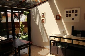 Ein Zimmer mit einem Holztisch, einem Stuhl, einem Schreibtisch mit Schubladen und einem kleinen Papierkorb. An der Wand hängen zwei gerahmte Bilder und ein Fenster mit Blick auf einen Garten.