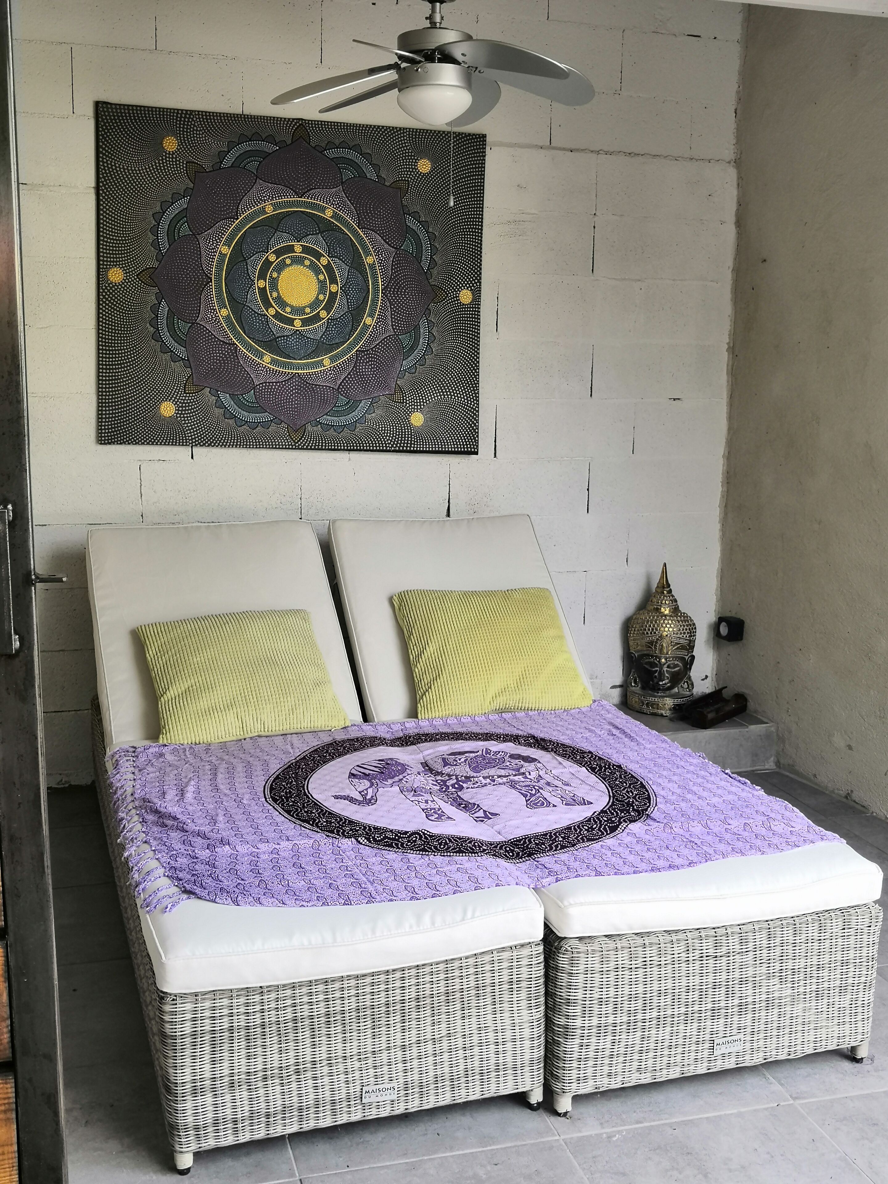 A bedroom with a bed, two pillows, a wall tapestry, a ceiling fan, and a Buddha statue.