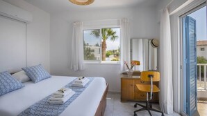 A hotel room with a bed, a desk, a chair, a window with a view of palm trees, and a balcony.