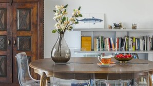 A dining area with a round table, clear chairs, a vase with flowers, a bowl of fruit, and a bookshelf in the background.