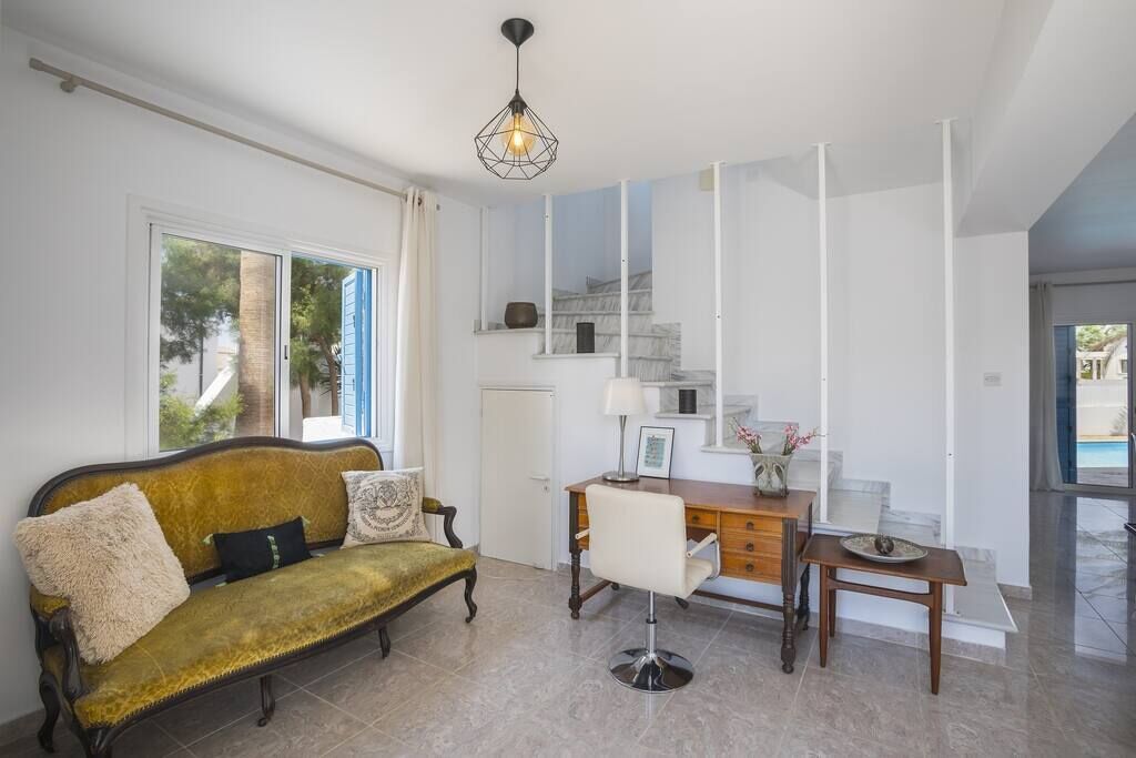 Una sala moderna con un sofá, un escritorio y una silla. Se ve una escalera con barandilla y vistas al exterior.
