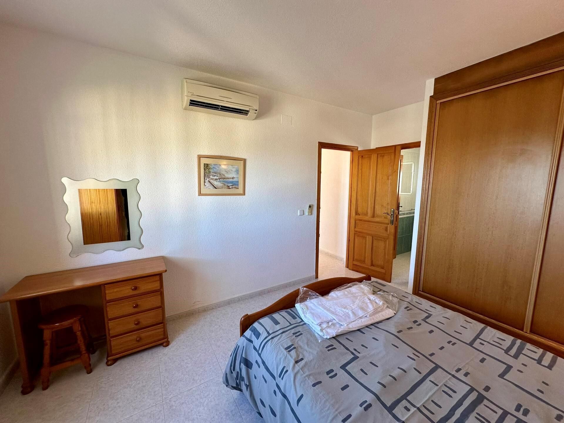 A bedroom with a bed, a wooden desk, a dresser, a framed picture, an air conditioner, and a wooden wardrobe.