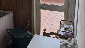 A room with a wooden interior, a white table, a green trash bin, a chair, and a view of a red brick wall and greenery outside. - Penthouse Anna, in the center of a small town with a splendid sea view (Ortona)