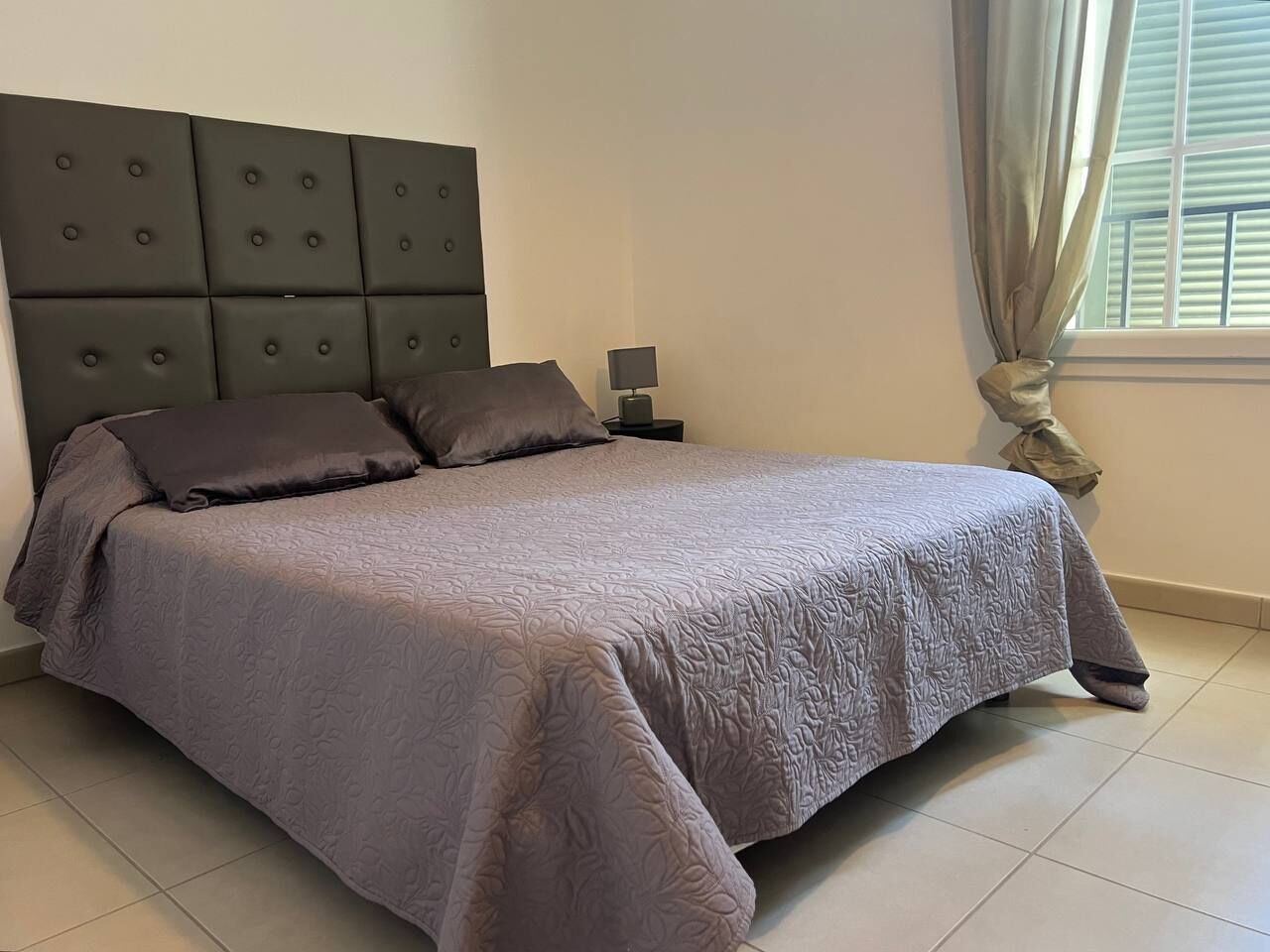 Een netjes opgemaakt bed met een grijze deken en bijpassende kussens, een donker hoofdbord, een nachtkastje met een lamp en een raam met gordijnen.