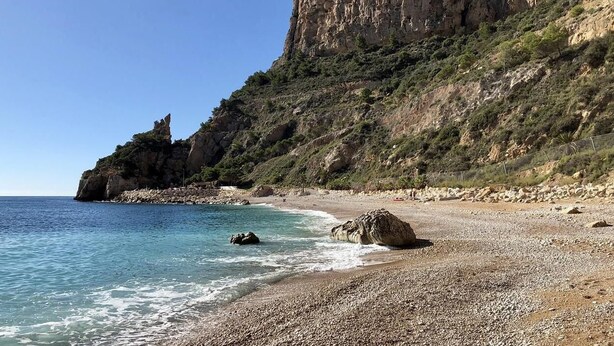 Une plage rocheuse bordée d’une eau d’un bleu limpide, une grande formation rocheuse et une falaise abrupte.